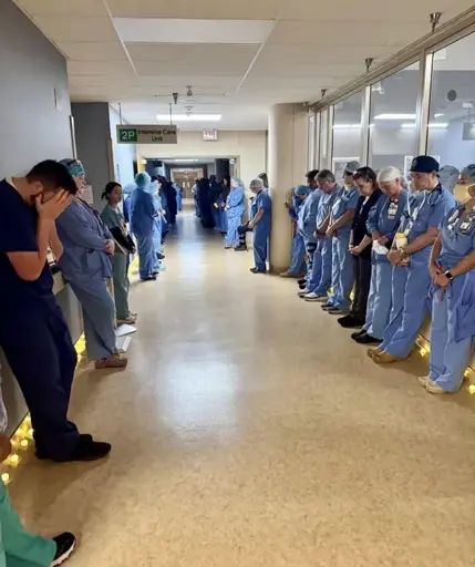 Nurses and doctors line the hallways of a hospital with bowed heads. One has their their head in their hands.