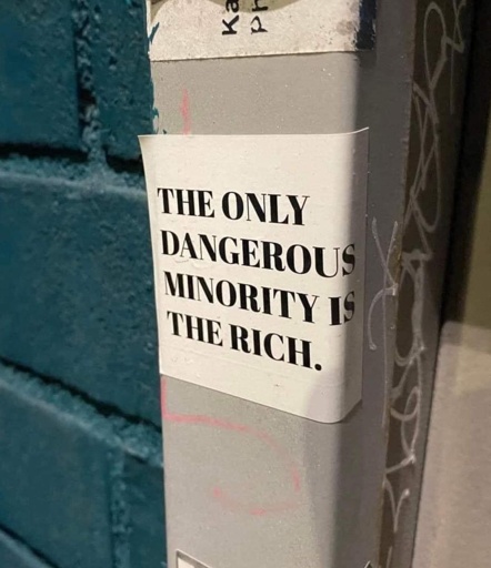 A street sticker pasted on a grey pole with graffiti around it reads in bold black capital letters: "THE ONLY DANGEROUS MINORITY IS THE RICH." The sticker contrasts against a teal brick wall in the background, making the political message stand out.