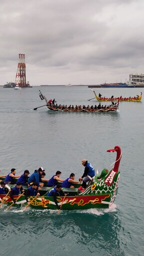 3 Hari boats racing, each with about 20 men paddling in the middle of a port with big ships in the background.