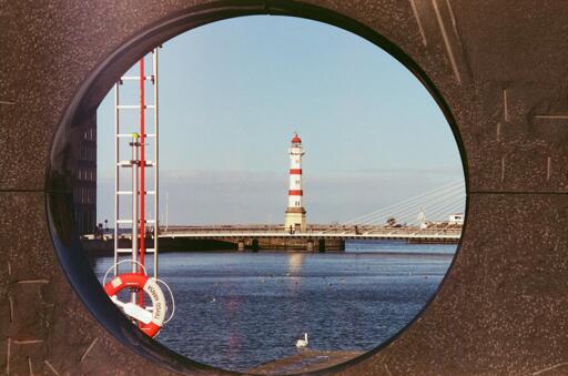A lighthouse and bridge are framed through a large circular opening in a wall, with calm water, a swan, and a red-white pole with a buoy in the foreground.