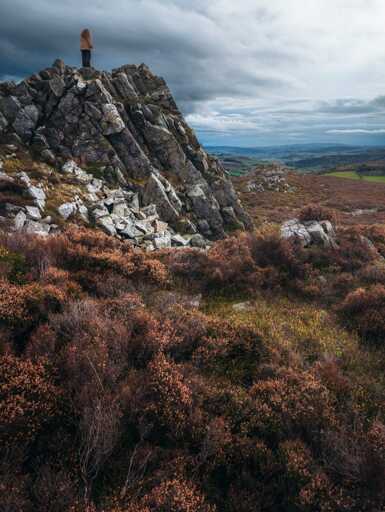 A lone person stands on top of a jagged outcrop of grey rock at Cranberry Rock on the Stiperstones, looking out over a broad rural landscape under a heavy sky. The crags rise sharply from heather moorland that fills the foreground with russet and earthy tones. Beyond the main ridge, rolling hills and patchwork fields stretch into the distance with muted autumn colours. Low cloud and soft, diffused light give the scene a moody and atmospheric feel.
